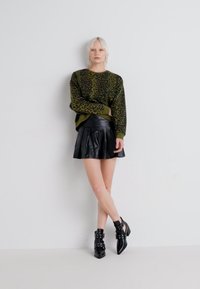 Maglione verde con stampa leopardata, gonna plissettata nera in faux pelle e stivaletti neri con dettagli in cinturino. Texture morbide e design aderente.
