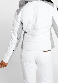 Vit och ljusgrå vadderad jacka med en figursydd design, med en slät textur och sidofickor med dragkedja. Matchad med vita byxor.