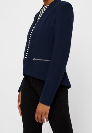 Kobieta nosząca dopasowaną marynarkę w kolorze granatowym z poziomą kieszenią na zamek oraz czarno-białą koszulę w paski pod spodem.