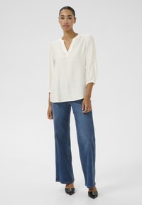 Blouse blanche légère avec un décolleté en v et des manches bouffantes trois-quarts, associée à un jean en denim bleu à jambes larges et des chaussures noires à bout pointu.