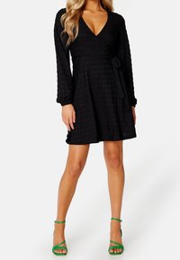 Schwarzes Wickelkleid mit langen Ärmeln, strukturiertem Stoff, V-Ausschnitt und gebundenem Taillengürtel. Kombiniert mit grünen Riemchen-Highheels.