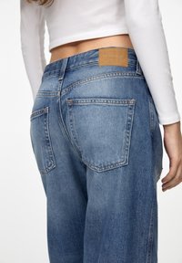 Světle modré denimové džíny s uvolněným střihem, které mají dvě zadní kapsy, koženou nášivku v barvě tan na pasu a viditelný detail šití.