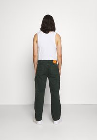 Zelené bavlněné cargo kalhoty s rovným střihem, s kapsami na zadní straně a oranžovou štítkovou nášivkou poblíž pasu, doplněné bílým tílkem.