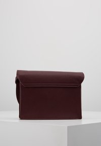 Borsa a mano in pelle bordeaux con superficie strutturata, patta pieghevole e forma rigida. Cuciture robuste visibili lungo i bordi.