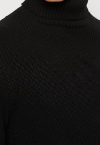 Jersey de cuello alto negro fabricado en punto acanalado, con un alto cuello y una superficie texturizada. Diseño duradero y ajustado.