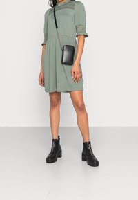 Femme portant une robe courte vert sauge ornée de dentelle, des bottines à talons épais noires avec des anneaux de fermeture éclair argentés, et un sac bandoulière noir.