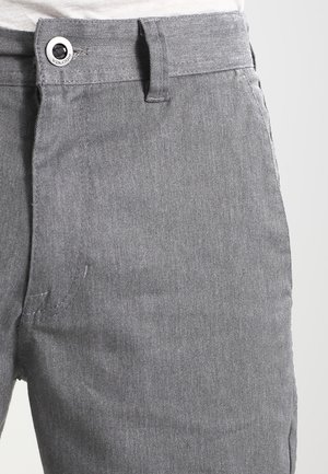 Gros plan d’un pantalon gris avec un bouton et une passants de ceinture à la taille, montrant la texture du tissu et les coutures de poche sur fond blanc.