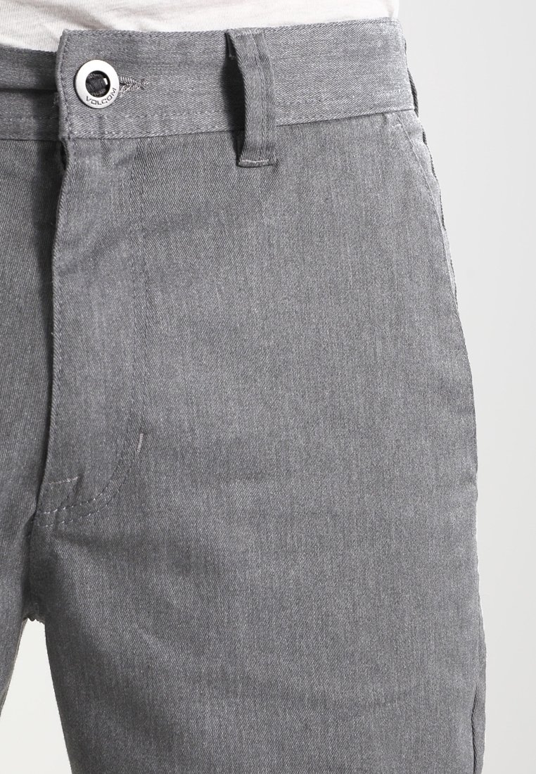 Gros plan d’un pantalon gris avec un bouton et une passants de ceinture à la taille, montrant la texture du tissu et les coutures de poche sur fond blanc.