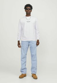 Long-sleeve white T-shirt with black text, light blue denim jeans, and tan boots, complemented by black sunglasses and a silver chain.