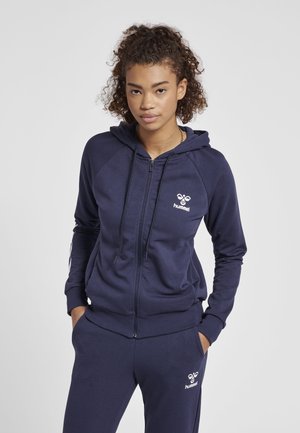 Granatowy dres sportowy z kapturem, z zamkiem błyskawicznym i raglanowymi rękawami. Posiada logo Hummel na piersi oraz białe akcenty. Miękka, teksturowana tkanina.