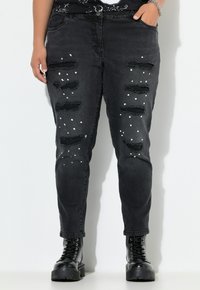Zwarte verwoeste jeans met meerdere scheuren, witte verfspetters, een taps toelopende pasvorm en rafelige zoom, gedragen met zwarte veterschoenen.