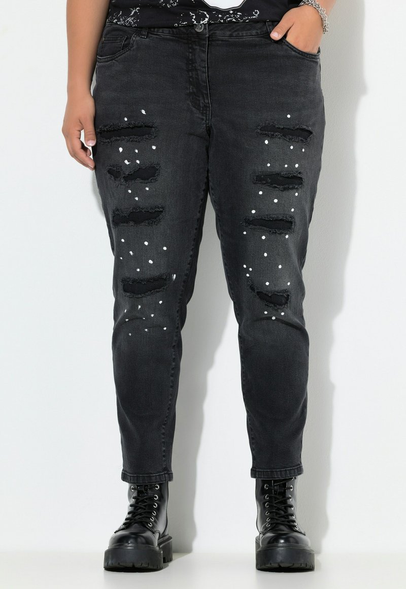 Zwarte verwoeste jeans met meerdere scheuren, witte verfspetters, een taps toelopende pasvorm en rafelige zoom, gedragen met zwarte veterschoenen.