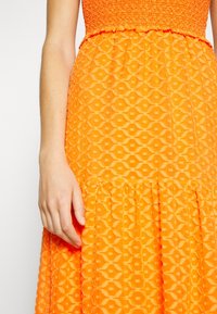 Gros plan d'une personne portant une robe orange vif avec une jupe texturée et à motifs, et un corsage style crochet, main détendue sur le côté.