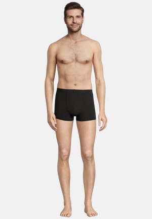 Boxers en coton noir ajustés, avec une ceinture texturée et un pouch frontale. Le modèle se tient pieds nus sur un fond neutre.