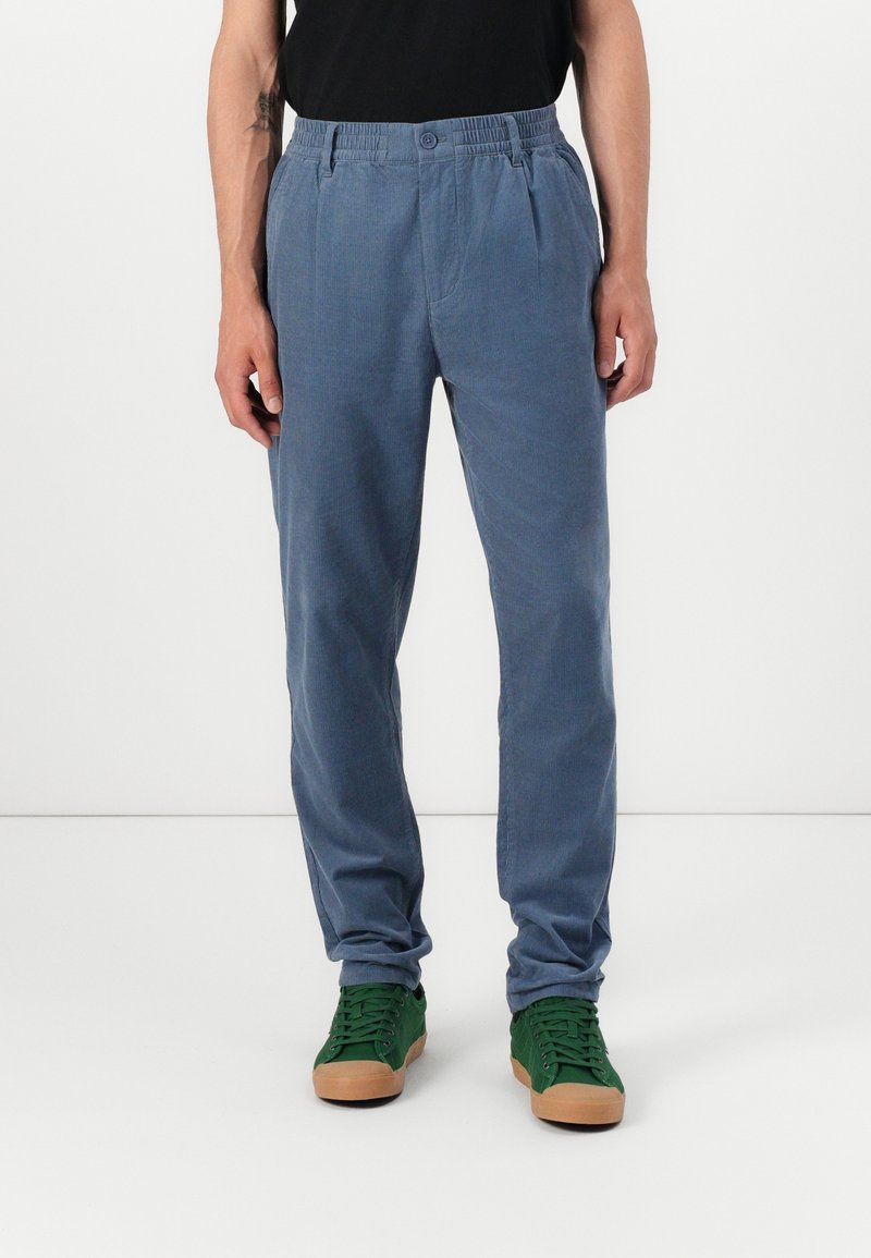 Personne portant un pantalon bleu droit et des baskets vertes avec des semelles marron, debout devant un fond blanc uni.