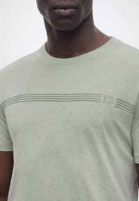 Άνδρας που φοράει ανοιχτό πράσινο μπλουζάκι με οριζόντιες ρίγες και ένα διακριτικό λογότυπο στο στήθος, μπροστά σε απλό φόντο.