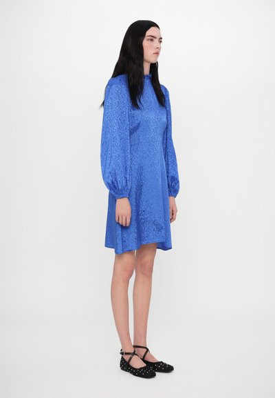Blauwe korte jurk met lange pofmouwen, een gestructureerd patroon en een gerimpelde kraag. Draagt bijpassende zwarte ballerina's met een bandje en stippen.