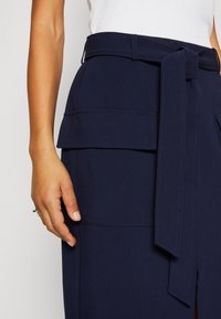 Gros plan d'une personne portant une jupe bleu marine avec une ceinture nouée et de grandes poches latérales, associée à un haut blanc sans manches.