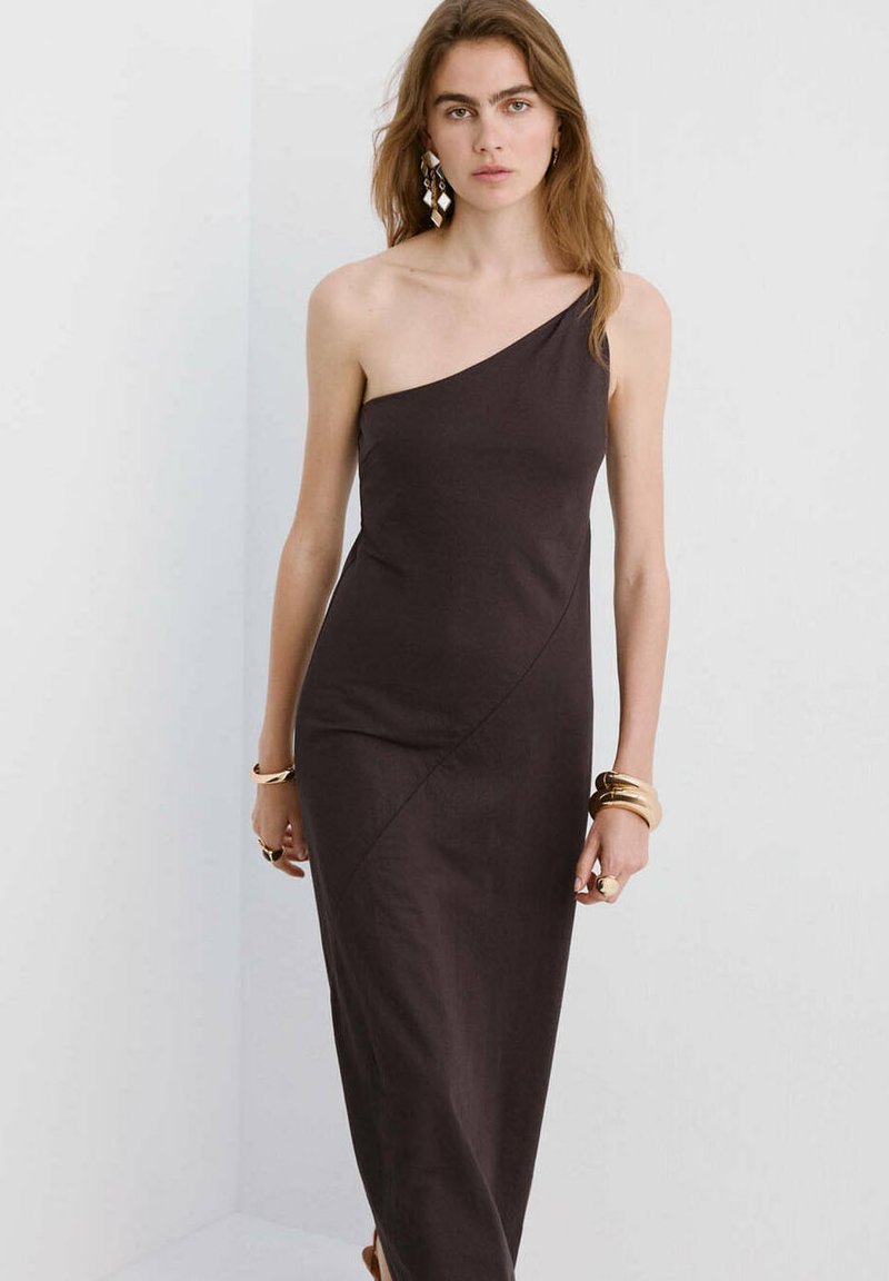 Femme portant une robe noire asymétrique sans manches avec des bracelets dorés et des boucles d’oreilles géométriques sur un fond blanc uni.