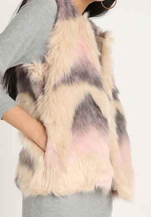 Vrouw draagt een mouwloos nepbontvest in crèmekleurige, roze en grijstinten over een grijze top met lange mouwen, met één hand in de vestzak.