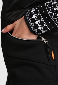 Giacca nera con una tasca con zip. Le maniche presentano un motivo geometrico bianco. Il materiale appare liscio con una leggera lucentezza.