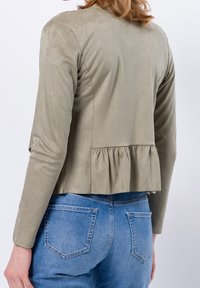 Giacca finta suède verde oliva con maniche lunghe e design a peplum, indossata su jeans in denim azzurro chiaro con due tasche posteriori.