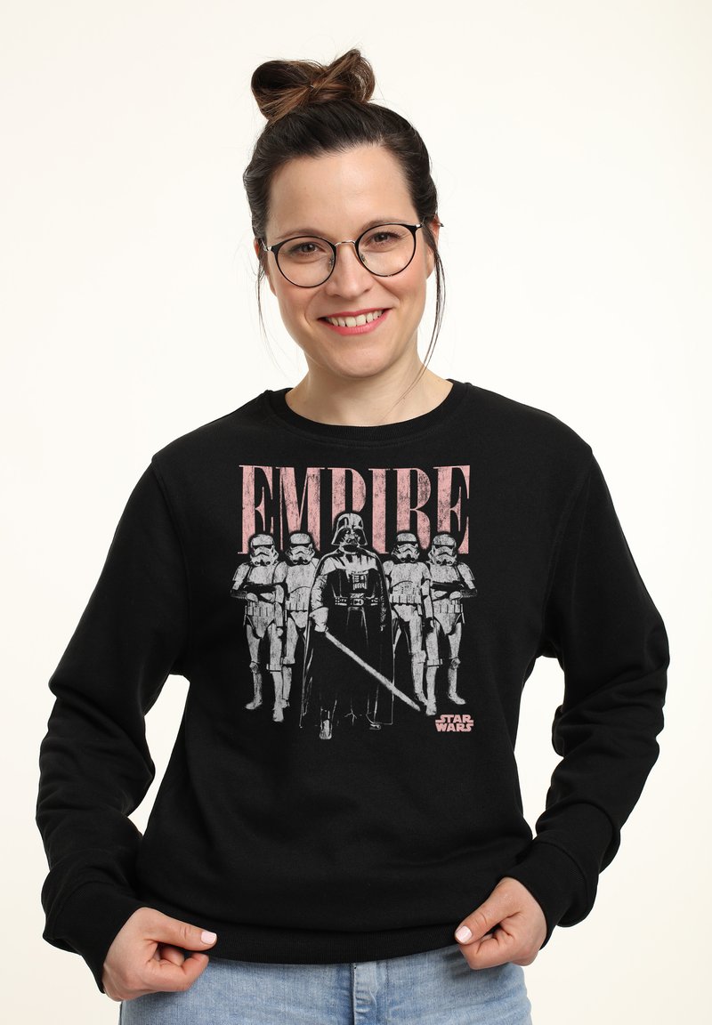 Smilende kvinde med briller iført en sort Star Wars-sweatshirt med Darth Vader og stormtroopers under ordet "EMPIRE."