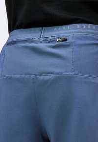 Gros plan sur un short de course Nike bleu avec poche arrière zippée et un haut noir visible à la taille.