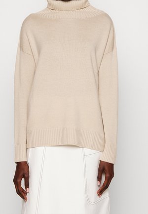 Pull à col roulé côtelé beige porté avec une jupe blanche présentant des coutures apparentes et un détail de poche.