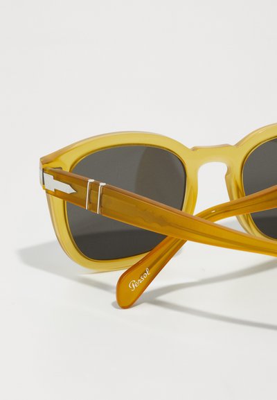 Um par de óculos de sol âmbar com lentes pretas, apresentando armações grossas em acetato e hastes leves com um destaque prateado e a marca "Persol".