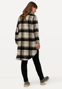 Kariertes Oberbekleidungsstück in Creme-, Schwarz- und Orangetönen, mit einem langen, geraden Design und einem asymmetrischen Saum. Trägt es über schwarzen Hosen und grauen Stiefeln.