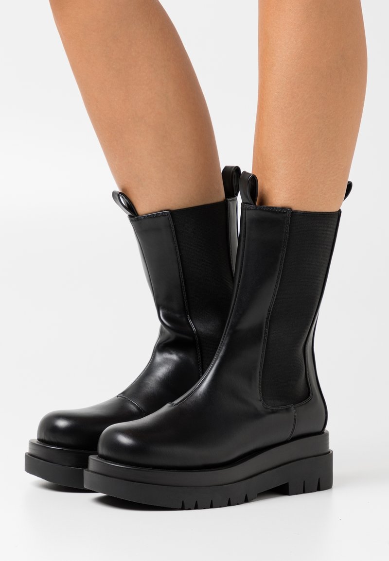 Bottes chevilles en cuir noir avec panneaux latéraux élastiques, semelles plateformes épaisses noires et languettes bouclées en haut pour un enfilage facile.