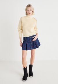 Pull en maille côtelée crème à col roulé, jupe plissée en denim bleu marine avec broderies florales rouges, et bottines noires avec accents métalliques.