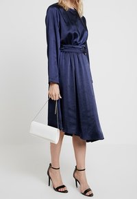 Abito in raso blu navy con maniche lunghe, vita con cintura e lunghezza midi; abbinato a una borsa bianca e tacchi neri con cinturini.