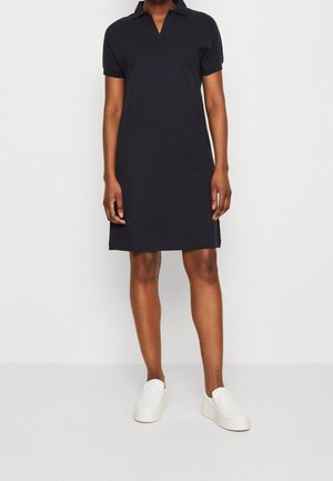 Marineblaues Polokleid mit kurzen Ärmeln, Kragen und geradem Schnitt. Getragen mit weißen Slippern. Glattes Material ohne sichtbare Muster oder Akzente.