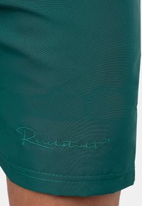 Pantaloni corti di un verde scuro realizzati in tessuto leggero, con testo ricamato "Reichstadt" dello stesso colore del filo lungo l'orlo.