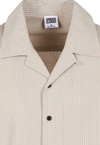 Beiges Kurzarmhemd mit strukturierten vertikalen Streifen, zwei schwarzen Knöpfen, einer Brusttasche und einem Kragen. Das Etikett trägt die Aufschrift "Urban Classics M."