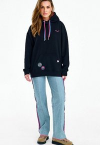 Žena v černé mikině s kapucí s barevnými šňůrkami a nášivkami, světle modrých kalhotách s růžovými proužky po stranách a béžových botách na šněrování.