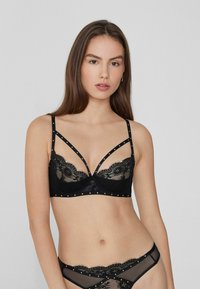Bralette de encaje negro con paneles transparentes, herrajes decorativos, tirantes ajustables y un intrincado detalle de encaje en el escote.