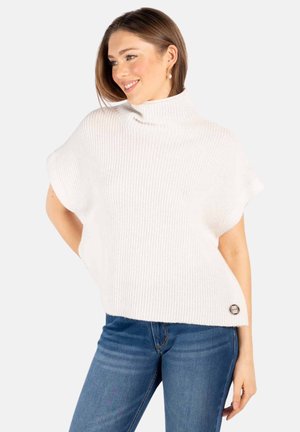 Kvinde med lysebrunt hår iført perleøreringe, en hvid kortærmet rullekravetrøje og blå jeans, smilende og kigger mod venstre.