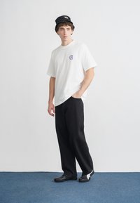 Biała koszula z krótkim rękawem z małym fioletowym logo, luźne czarne spodnie i czarny kapelusz typu bucket. Czarne obuwie z białymi akcentami.