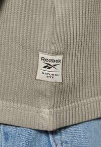 Grå ribbad textil med en Reebok-logetikett som läser "NATURAL DYE." Fokuserad på tygtextur och etikettens detaljer nära denim.