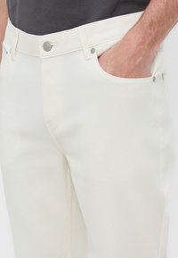 Homme portant un pantalon écru avec la main dans la poche avant et un bouton argenté visible ainsi que des rivets à la taille et aux poches.