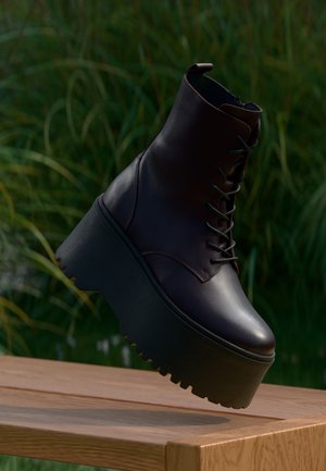 Dark brown platform ankle boot with thick ridged sole and laces, shown floating above a wooden bench with green foliage background.