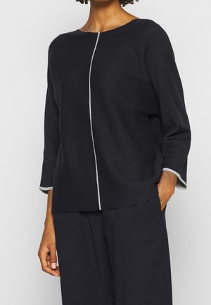 Femme portant un haut noir à manches longues avec une bande verticale blanche au centre et un pantalon noir assorti, main dans la poche.