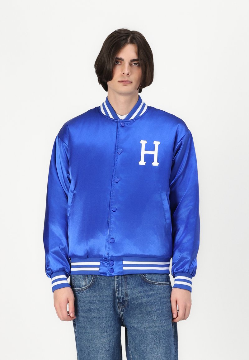 Jachetă de varsity din satin albastru cu logo-ul alb "H", guler și manșete stripe, precum și butoane frontale și buzunare laterale. Stilizată cu blugi din denim deschis.