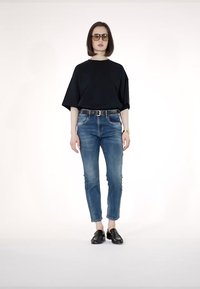 Črna oversize majica, modri oprijeti kavbojke in črne slip-on čevlje. Model nosi velike sončne očale. Enostavna bela podlaga.