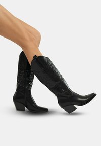 Botas negras hasta la rodilla con punta afilada, superposición texturizada, cremallera lateral y un tacón bajo en bloque; presenta patrones en relieve de espirales.