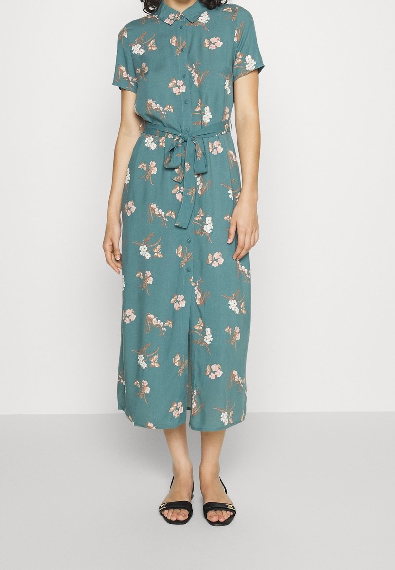 Robe midi bleu sarcelle à imprimé floral rose et blanc, manches courtes, boutonnée sur le devant, ceinture à la taille, portée avec des sandales noires à bouts ouverts.