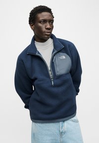 Άνδρας με ναυτικό μπλε φλις μπουφάν με τσέπη στο στήθος και ανοιχτό μπλε τζιν, κοιτάζοντας πλάγια σε απλό φόντο.